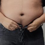 Hombre con obesidad intenta abrocharse el pantalón pero no lo logra porque su abdomen en muy grande y tiene exceso de peso